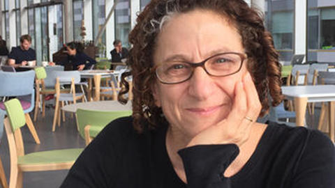 Cynthia Dwork, a light-skinned woman with curly auburn hair and glasses, sits at a cafe in front of a bank of windows.