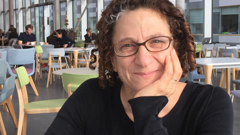 Cynthia Dwork, a light-skinned woman with curly auburn hair and glasses, sits at a cafe in front of a bank of windows.