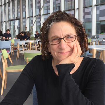 Cynthia Dwork, a light-skinned woman with curly auburn hair and glasses, sits at a cafe in front of a bank of windows.