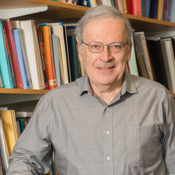 Leslie Valiant. Headshot. Les is facing the viewer. Behind him is a bookshelf full of many brightly-colored books. He has short white hair and glasses and is smiling.