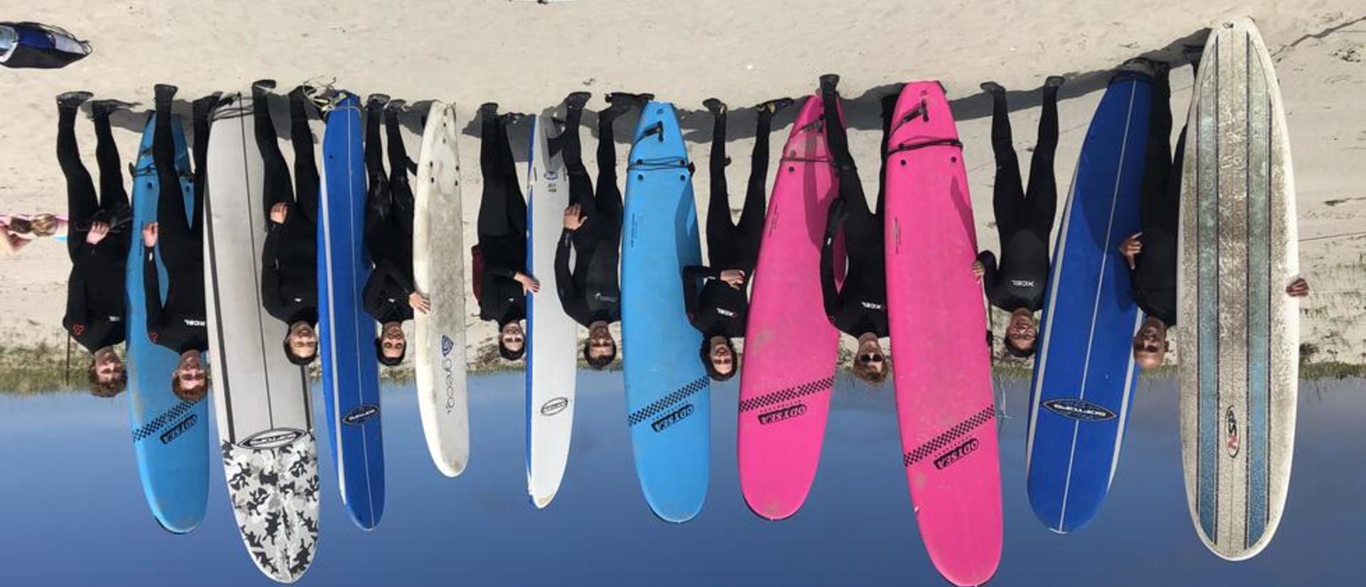 Ten smiling people pose with a variety of surfboards on a sunny beach. Everyone is wearing a wetsuit, so the water must be cold!