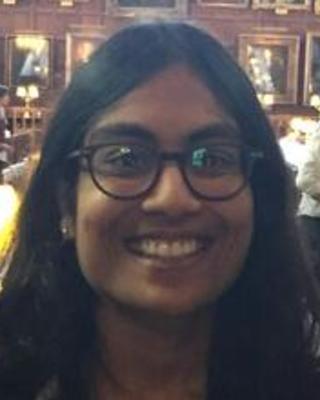 Mitali has brown skin and shoulder-length black hair. She is standing in front of a wall of pictures and smiling broadly at the camera. She wears black glasses with thick stylish rims.