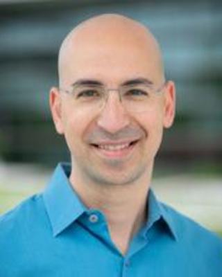 A front-facing headshot of Ariel. He has light skin, is bald, and is wearing glasses.