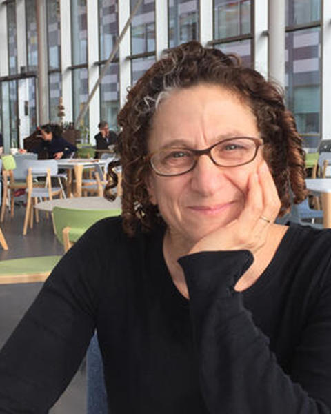 Cynthia Dwork, a light-skinned woman with curly auburn hair and glasses, sits at a cafe in front of a bank of windows.