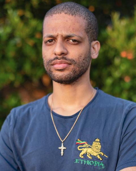 Jelani has medium-brown skin and dark eyes, with very short/ shaved black hair and beard. He is standing looking at the camera with his arms folded.