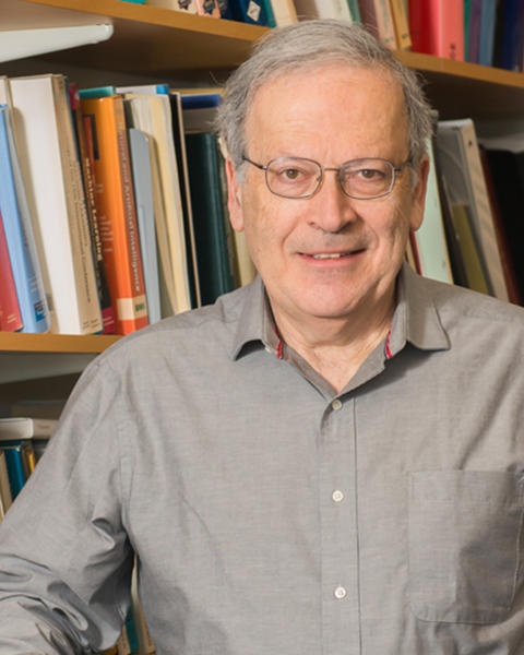 Leslie Valiant. Headshot. Les is facing the viewer. Behind him is a bookshelf full of many brightly-colored books. He has short white hair and glasses and is smiling.