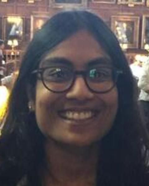 Mitali has brown skin and shoulder-length black hair. She is standing in front of a wall of pictures and smiling broadly at the camera. She wears black glasses with thick stylish rims.