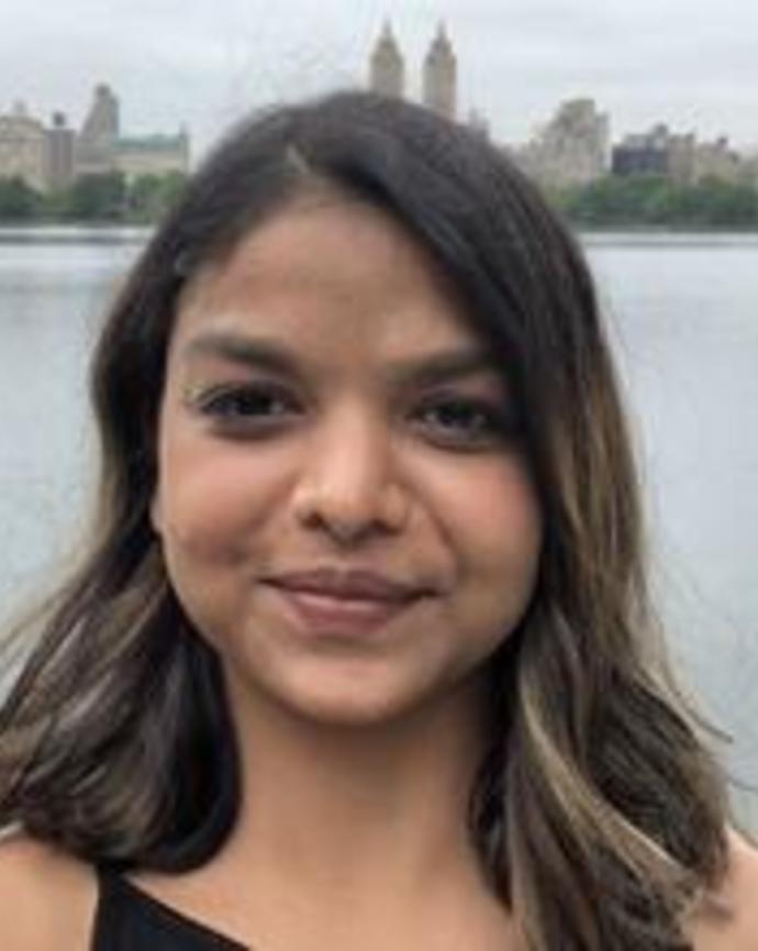 Yamini has dark olive skin and medium-brown wavy shoulder-length straight hair. She stands on a bridge in front of a cityscape in the photo.